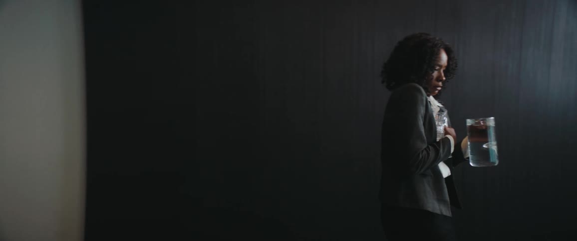 A woman in a gray blazer and white blouse carries two glass pitchers of water against a dark wall looking tense