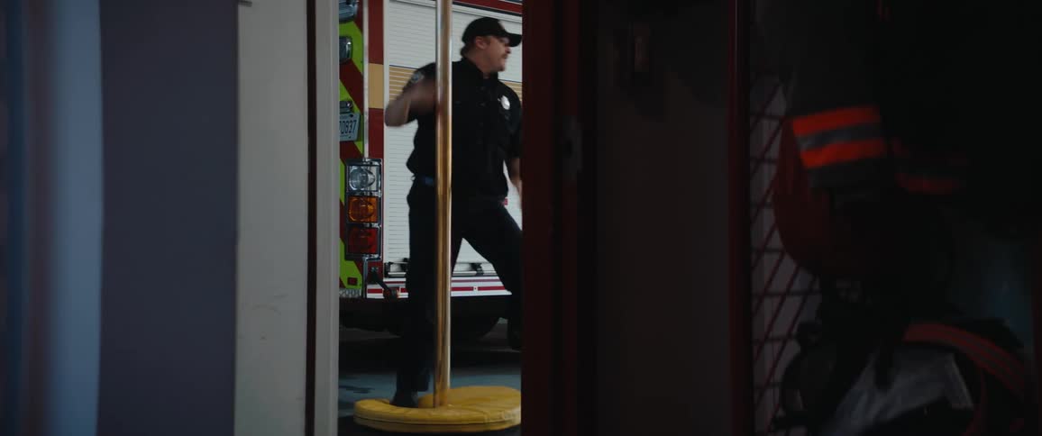 A male firefighter in a dark uniform rapidly slides down a brass fire pole in the interior of a fire station briefly stepping out of view as a large red and white fire truck is parked visible just outside the bay doors in the background