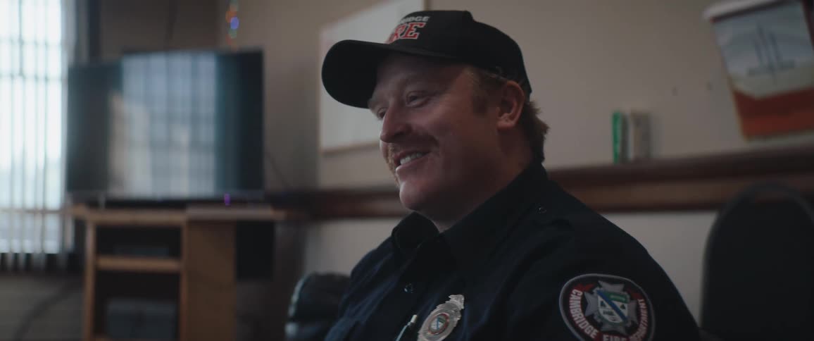 A man wearing a dark uniform shirt and a baseball cap sits and smiles while looking away from the camera in an indoor lounge area with a television screen in the background