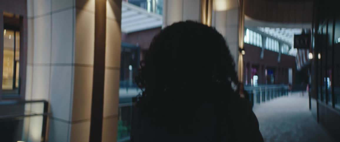A person with curly hair walks through a modern softly lit atrium at dusk while other commuters blur by on either side