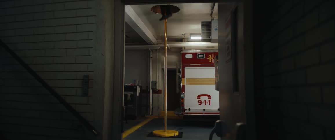 The video shows a person in dark clothing sliding down a fire pole in what appears to be a fire stationin the background visible through an open bay door a large red and white fire truck or ambulance with a 911 decal is parked
