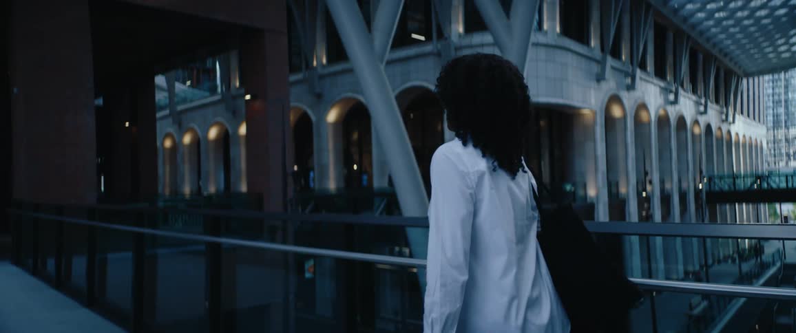 The video is an overtheshoulder shot of a black woman with curly hair walking across a modern pedestrian bridge or walkway at duskshe is wearing a white buttondown shirt a black bag is draped over her shoulder and the background features a