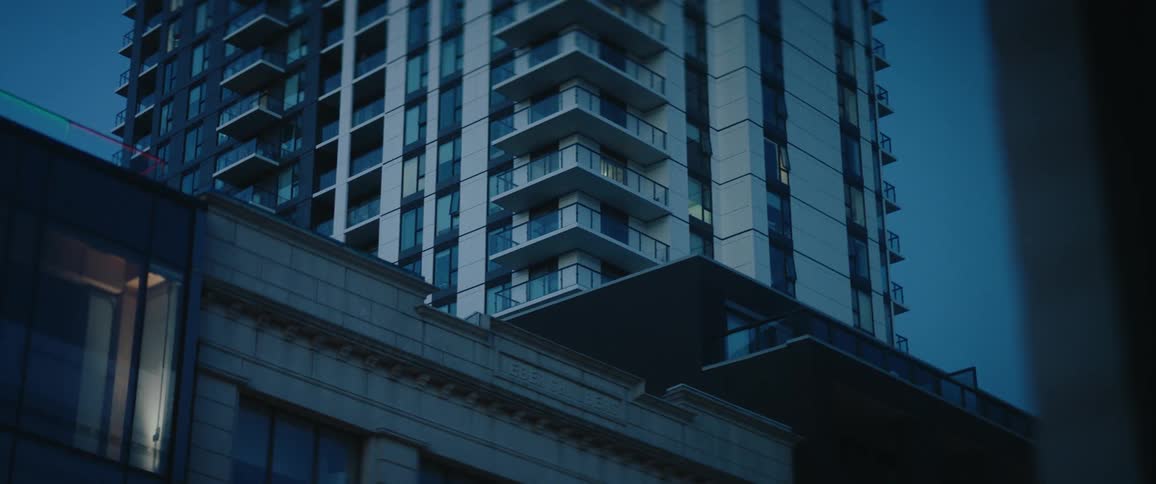 The scene captures the exterior of a modern highrise tower featuring many stories of balconies and reflective glass windows this structure sits atop the darker more traditional stonework facade of a shorter building below it