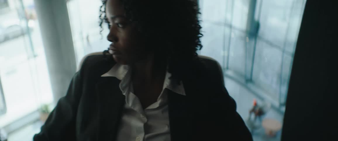 A young woman with curly dark hair wearing a white shirt and dark blazer is seen in profile near a large glass window looking down over the edge into the bright modern atrium of an office building with blurry figures visible far below