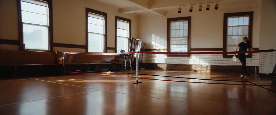 A man and a woman stand on opposite sides of a short net on a polished wood floor in a large sunlit room with multiple vertical windows and exchange hits during a badminton game