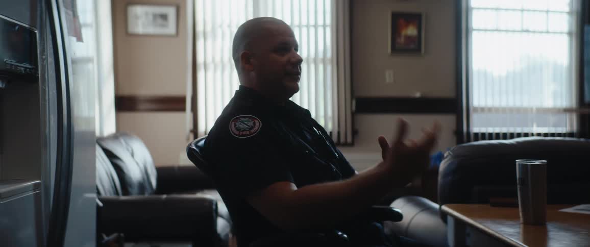 A middleaged man in a dark publicsafety uniform sits in profile on a lounge chair while talking and gesturing with his hands in a softly lit office with blinds