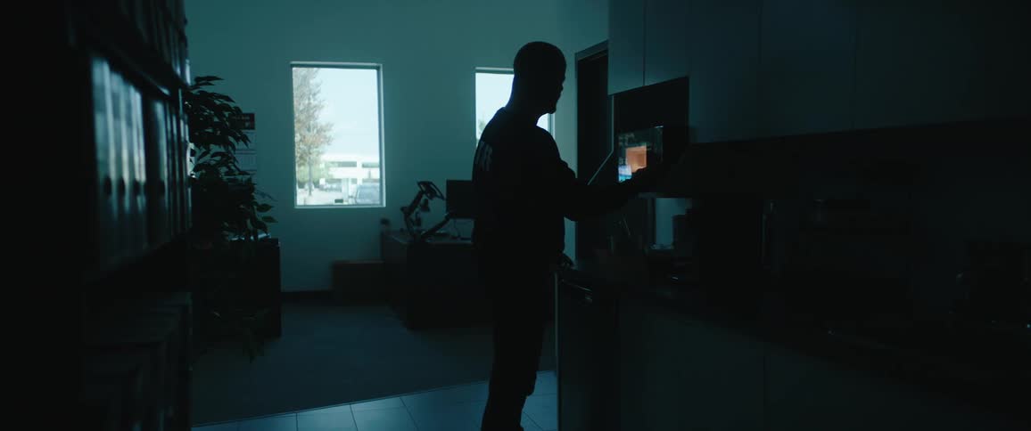 Bald man waiting in front of a microwave in a brightly lit office kitchen