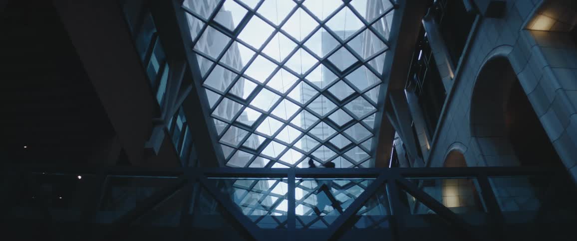 The view looks up into the ceiling of a modern multistory building where a glasspaneled atrium forms a striking diamondpatterned skylight a persons silhouette briefly passes across a lower pedestrian bridge visible through the glass str