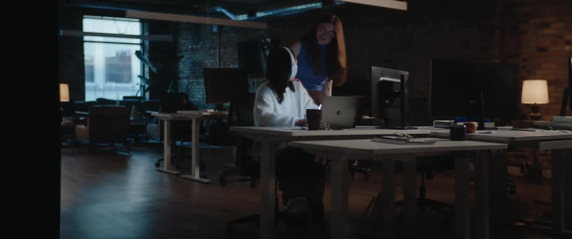 In a dim modern office at night a seated woman with headphones looks up from her laptop as a redhaired colleague leans in smiling both appearing engaged and friendly while collaborating