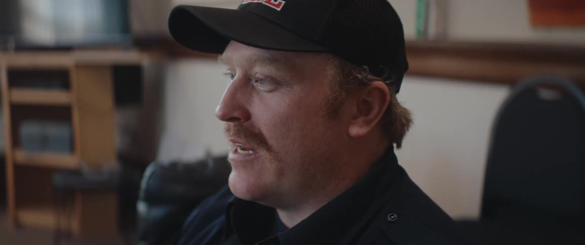 A medium closeup shows a man with reddishblond hair and a mustache wearing a black collared shirt and a matching baseball cap he is seated indoors speaking or listening with an engaged expression as he looks offcamera to the side