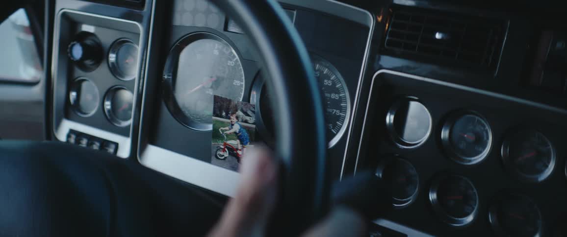 The interior dashboard of a truck is shown with a picture of a young boy on a small red bicycle taped over the face of one of the gauges a drivers hand partially visible in the foreground grips the black steering wheel