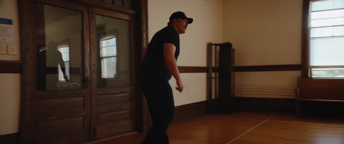 The video shows a man wearing a black cap and shirt who is holding a red white and blue ball while standing in the doorway of a room with wooden floorshe walks toward the right side of the frame and then turns to look back at the center of the ro