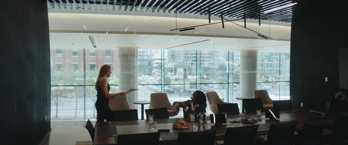 Woman hands another woman a folder in a large meeting room with glass windows