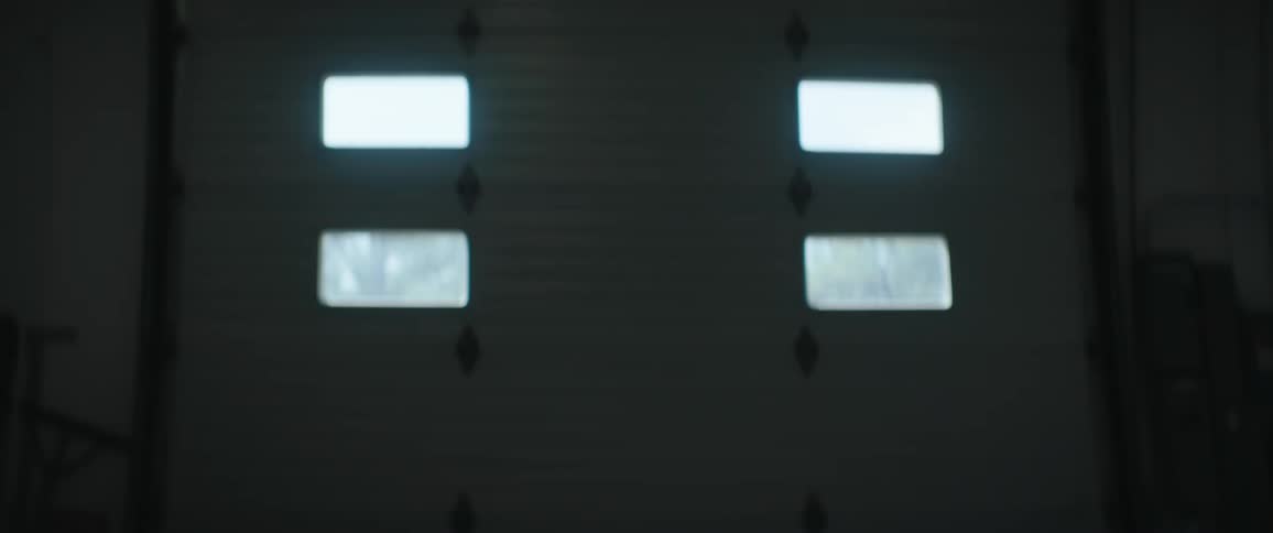 A bald man in silhouette is briefly seen walking in front of a closed garage door with four bright opaque window panels brushing his teeth before turning away into the darkness