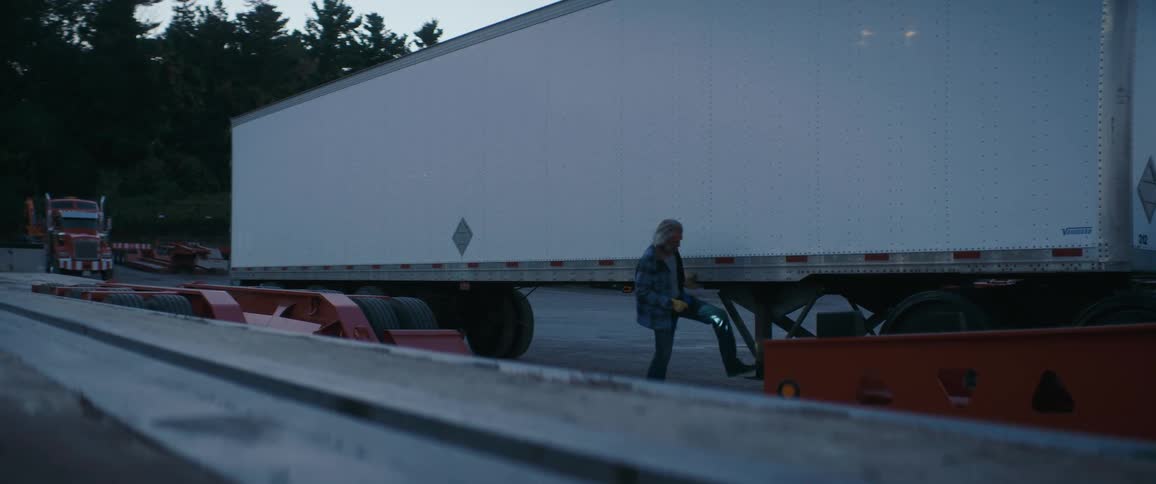 A truck driver in a plaid shirt and jeans is walking alongside a large white semitrailer in a parking or storage yard the driver stops and appears to be inspecting or adjusting something on the landing gear or frame of the trailer