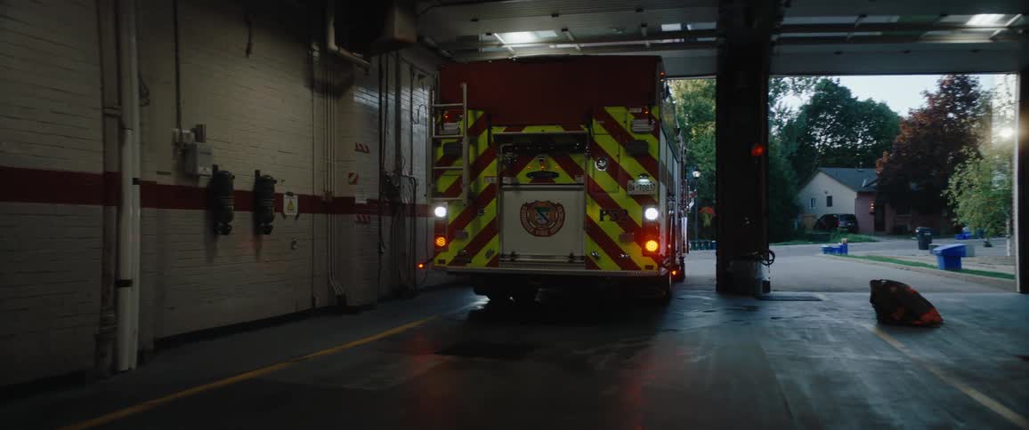 Inside a firestation bay a uniformed man gestures toward a departing fire truck with chevron striping the wet concrete floor reflecting the open doorway and evening light