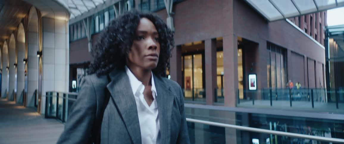 The video shows a mediumclose shot of a black woman in business attirea white shirt and a gray blazerwalking purposefully along an urban walkwayshe adjusts the collar of her shirt as she walks framed by the dark modern architecture of a ci