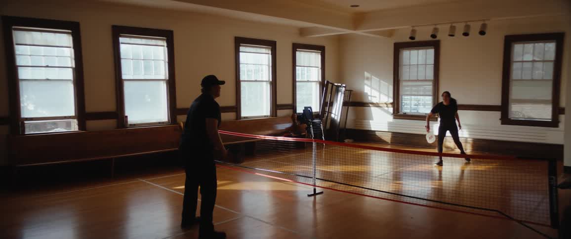 In a sunlit gym room two people in dark athletic wear rally across a portable net casually playing pickleball with an easy upbeat vibe