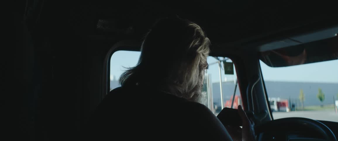 The back of a mans head with long light hair is seen in dark silhouette inside the cab of a truck as he holds a dark phone and turns slightly to look out the side window at a brightly lit industrial setting with blue sky and buildings