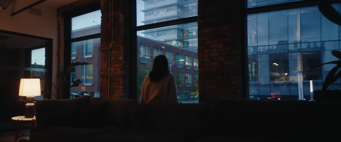 Woman standing before large windows observing illuminated buildings at dusk