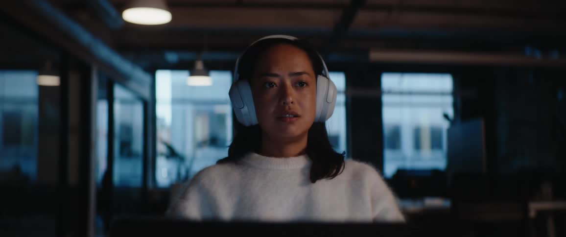 Young woman sitting down wearing headphones in front of a computer getting startled when another young woman taps her on the shoulder indoors