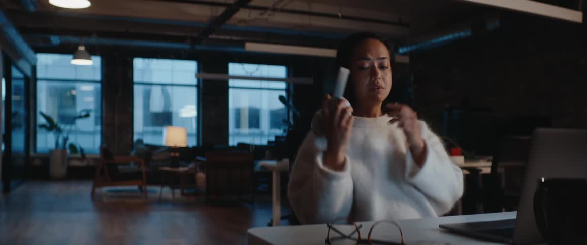 A woman in a white sweater and overear headphones focuses intently on her laptop at a dim office desk as a coworker walks past and waves
