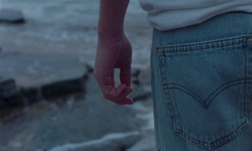 Person wearing a white shirt and jeans standing near the sea with focus on their hand and back pocket