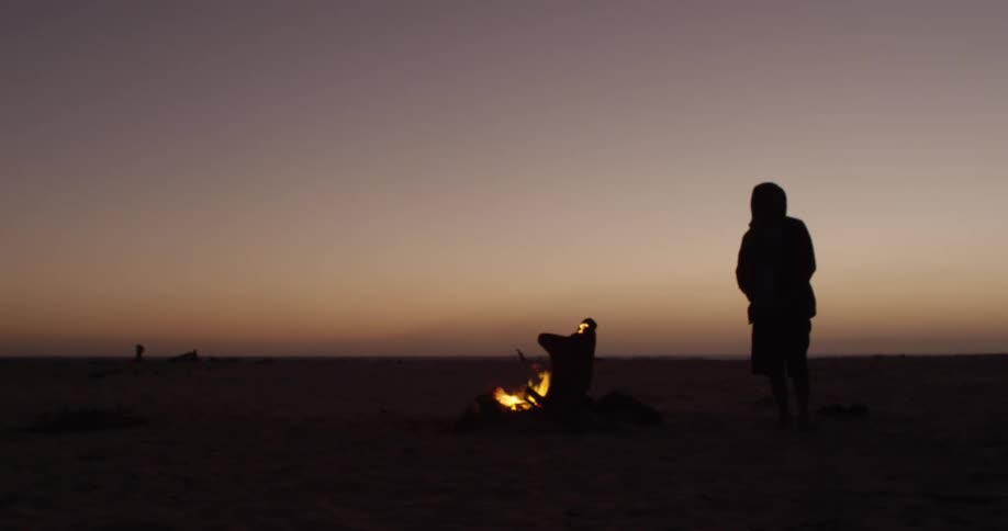 Person standing and watching a campfire burning on the beach at dusk