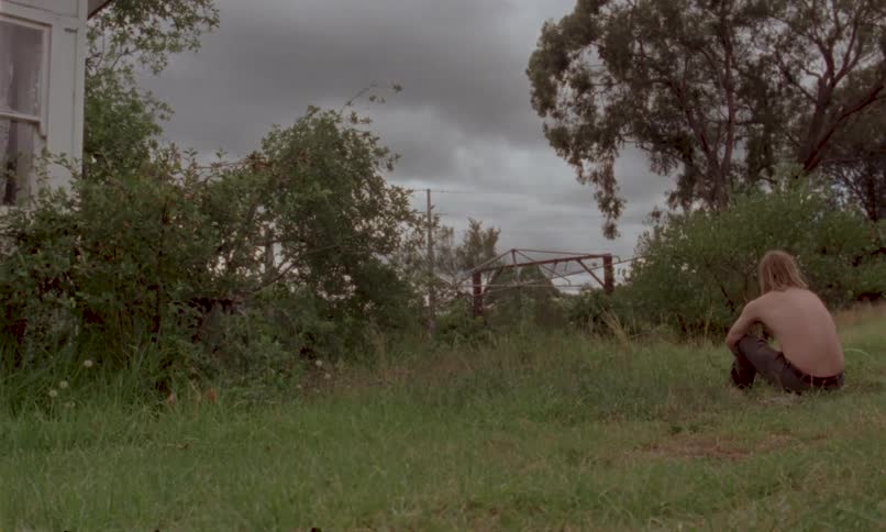 Shirtless person sitting on the grass beside a house under a cloudy sky