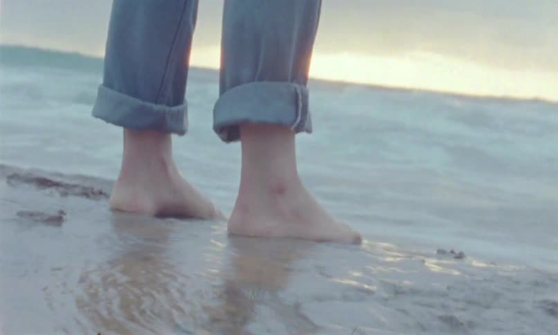 Bare feet on wet sand at the beach with rolledup jeans and gentle waves in the background