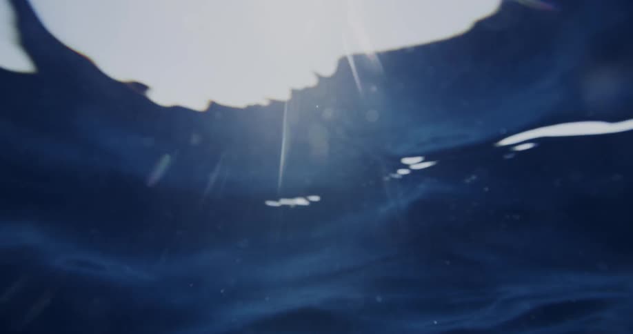 The video shows an underwater shot of deep blue water with the surface overhead bright light breaks through the waters surface creating sun rays and lens flares