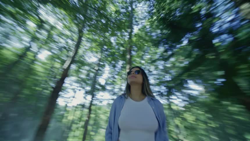 A woman in sunglasses looks up while standing in a sunlit forest