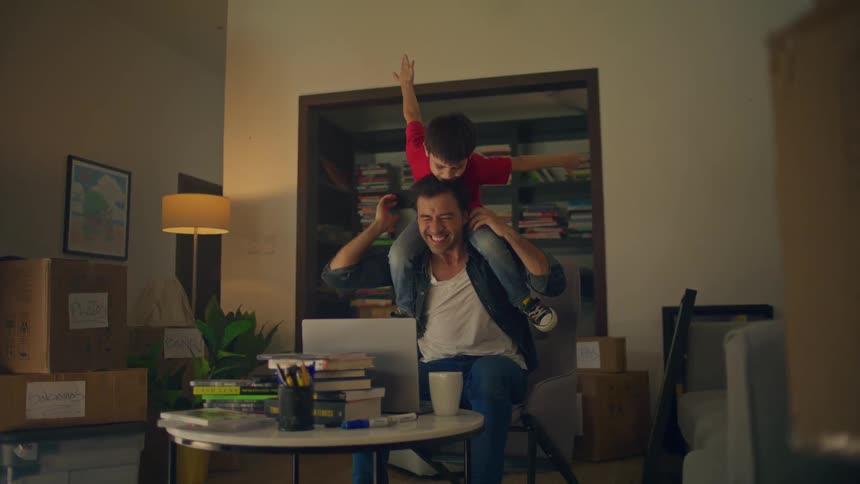 A man laughs with joy at a cluttered home desk as a little boy in a red shirt rides on his shoulders with arms outstretched like an airplane