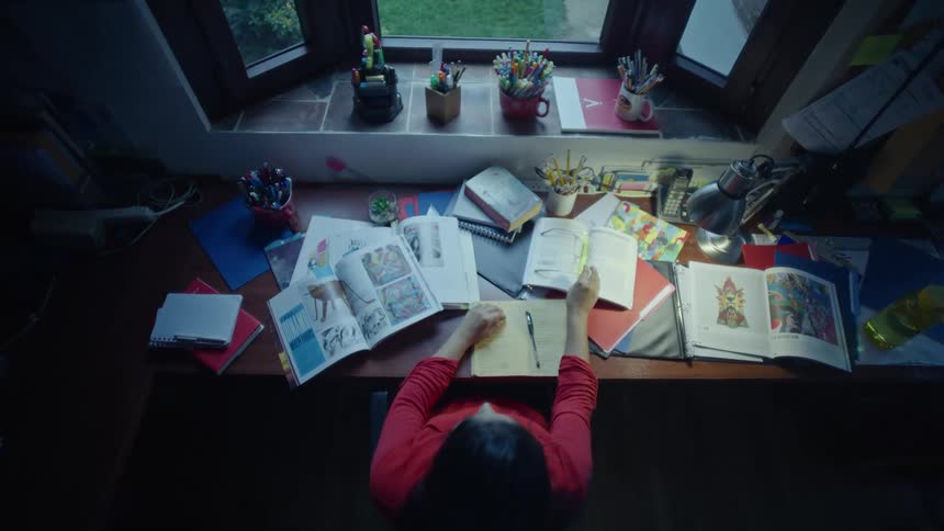 Person studying at a desk filled with books notebooks and colorful materials near a window