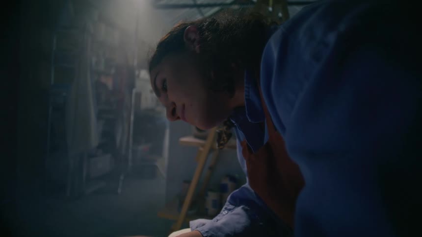 A young woman stands inside a workshop wearing a blue shirt and a red apron surrounded by tools and equipment