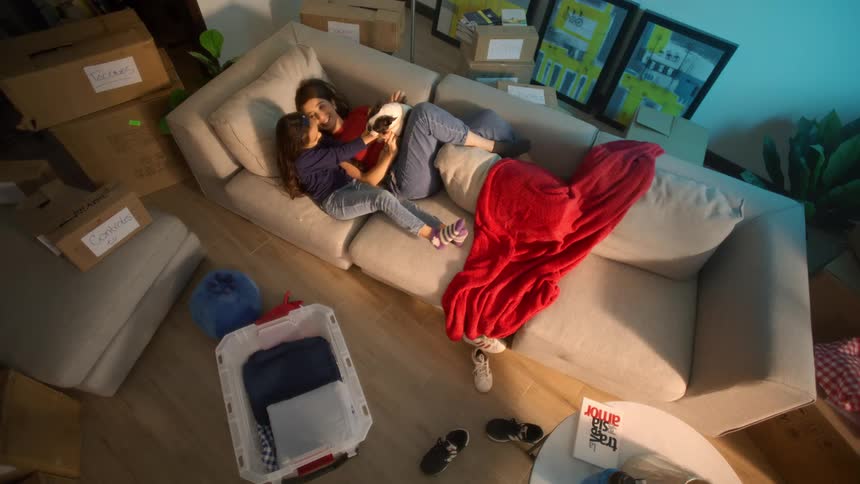 The video filmed from an overhead perspective shows a woman and a young girl lying comfortably on a couch covered in a red blanket they are nestled together looking at a small electronic device like a phone or tabletthey are surrounded by moving bo
