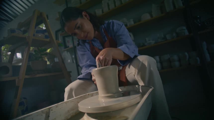 A young woman works inside a workshop at midday shaping a clay pot on a pottery wheel with focused hands and steady motion