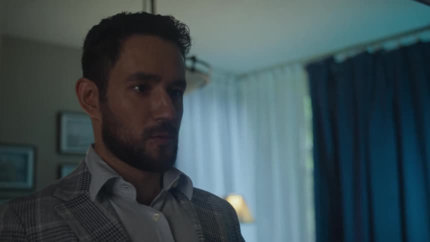 A well dressed man with a beard adjusts his plaid jacket while looking at his reflection in a mirror