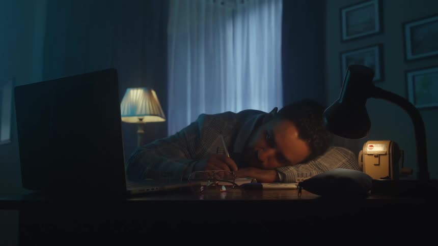 Man asleep on his desk beside a laptop and a lit desk lamp