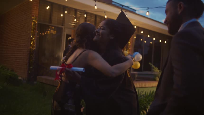 A hispanic woman wearing a graduation gown and cap stands on a grass lawn at dusk holding a diploma scroll and embraces another woman while a man stands nearby