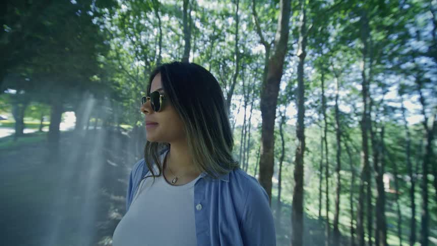 The video features a medium closeup of a woman wearing sunglasses a white shirt and a blue buttondown shirt outdoors she is standing in a lightly wooded area with tall trees and dappled sunlight shining through the leaves creating a bright slightly