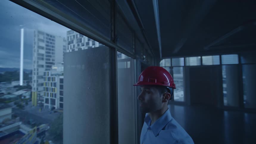 A man in a red hard hat looks out from a highrise building at a futuristic cityscape