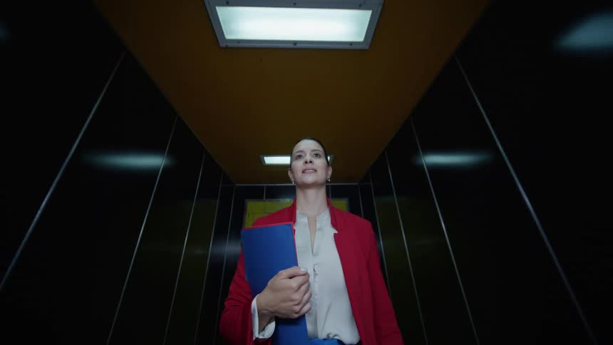 A woman in a red blazer walks in a modern building holding a blue folder to her chest and looking upward with a calm confident smile under the ceiling light