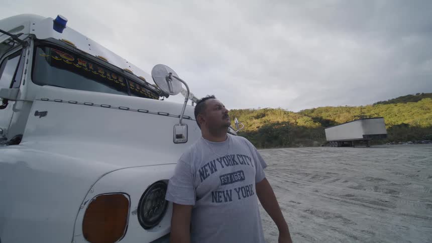 A middle aged hispanic man standing in front of a large cargo trailer parked outdoors during daytime