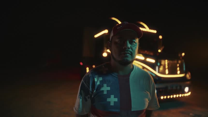A hispanic man wearing a cap and a patterned shirt stands in the darkness and looks at the camera while a large semitruck with its lights glowing sits directly behind him