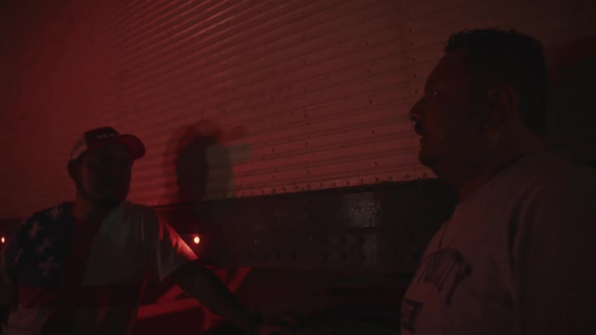 The video shows two men talking indoors bathed in a strong dark red light that gives the scene an intense and somewhat dramatic mood the man in the foreground is speaking partially obscuring his face from the cameras perspectivethe second man wea