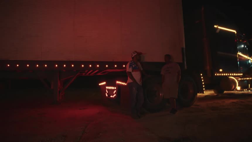 At night two men stand and talk next to a large semi truck which is illuminated by its running lights