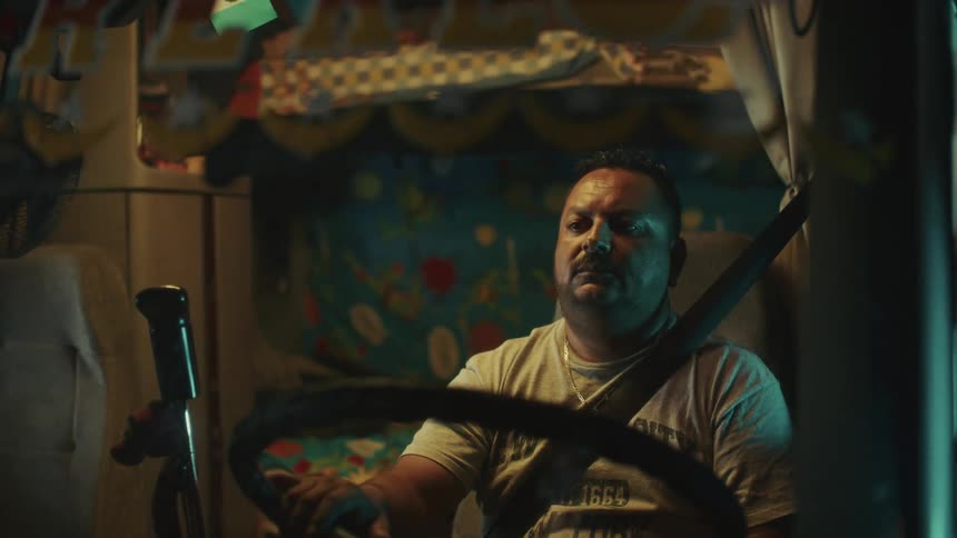 A hispanic man with beard sits in the drivers seat of a large vehicle holds the steering wheel and wears a shoulder seatbelt in a dark cabin decorated with fabric patterns