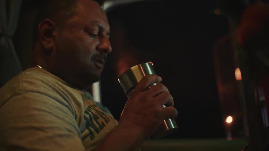 Close up of a middle aged man drinking a hot drink from a cup while sitting in a vehicle at night