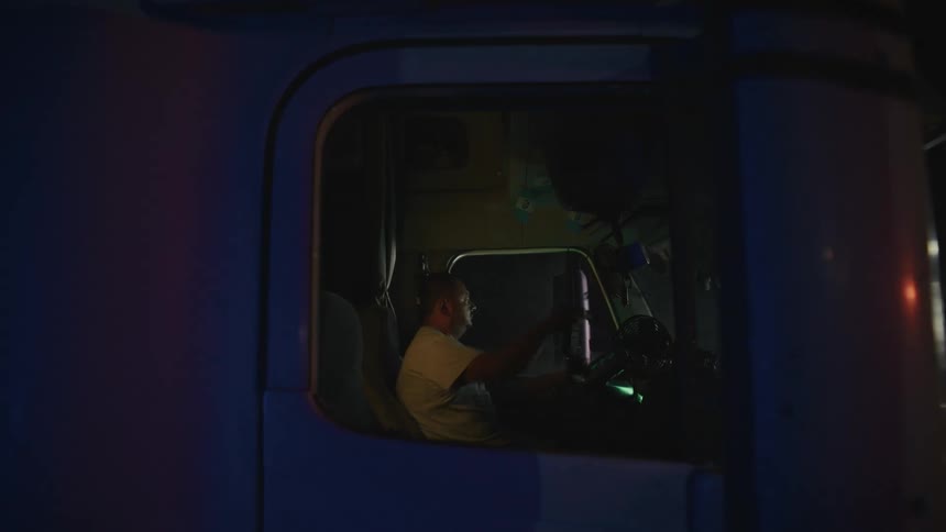 The video shows a man sitting in the cab of a large truck or semitrailer at night he is visible through the drivers side window illuminated primarily by the ambient light of the street and the vehicles dashboardthe man appears to be resting or wai